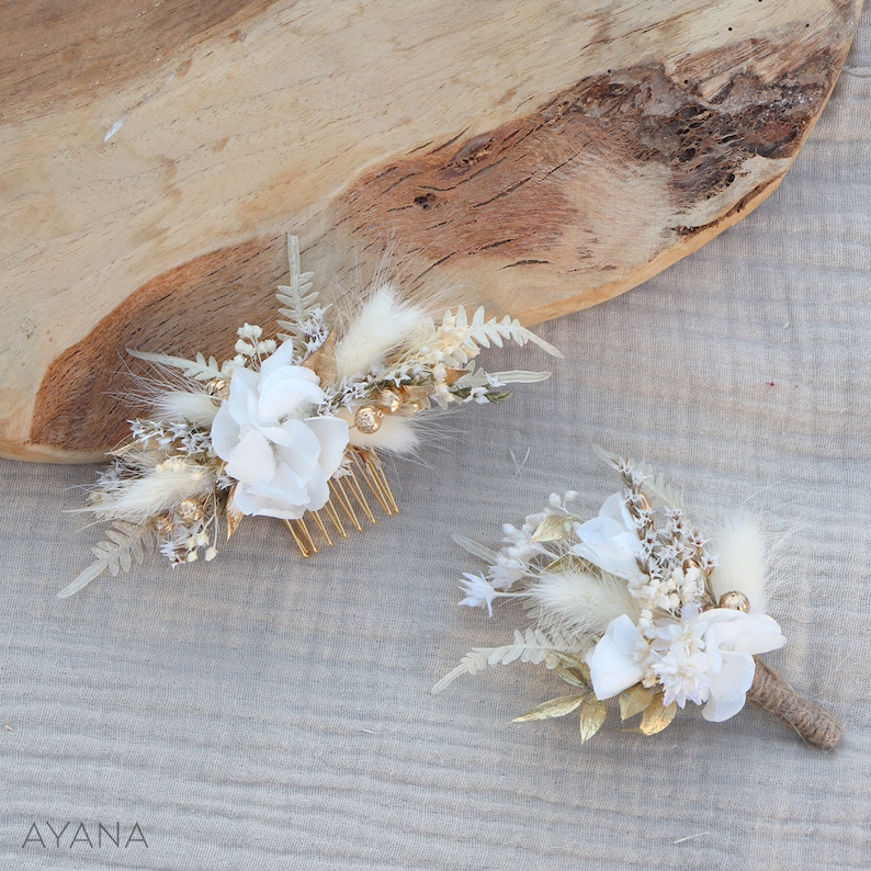 May include: Two white floral hair combs with gold accents. The larger comb has a white flower and the smaller comb has a white flower and a brown stem. Both combs are on a light brown fabric background.