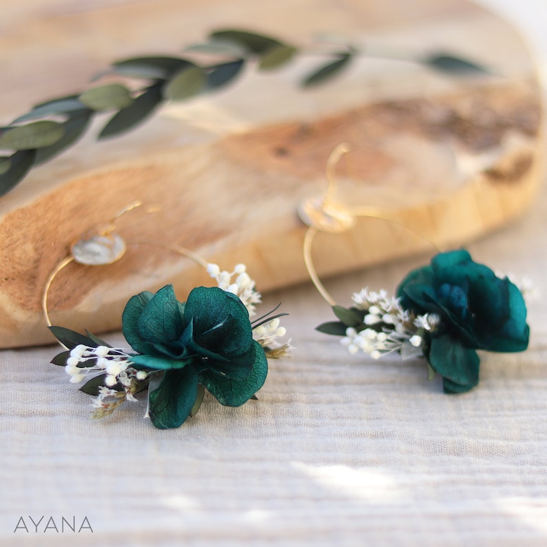 Op de afbeelding: Een paar gouden creolen met een turquoise bloem en witte gedroogde bloemen die aan elke creool zijn bevestigd.