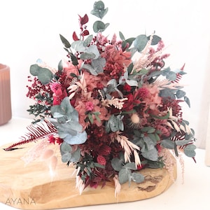 May include: A dried flower bouquet in shades of pink and burgundy with eucalyptus greenery. The bouquet is arranged on a wooden surface.