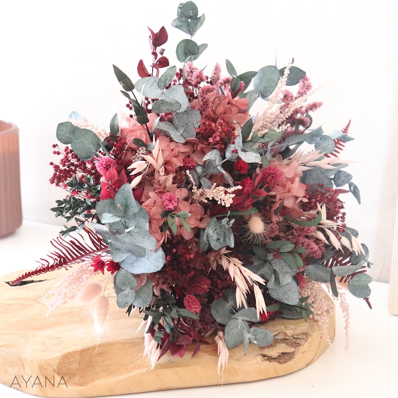 May include: A dried flower bouquet in shades of pink and burgundy with eucalyptus greenery. The bouquet is arranged on a wooden surface.