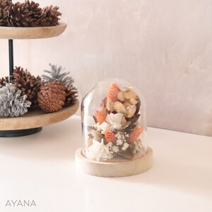 Op de afbeelding: Een glazen stolp met een houten voet die gedroogde bloemen bevat in tinten oranje, wit en bruin. De bloemen zijn in een tros gerangschikt en omvatten kleine witte bloemen, oranje bloemen en bruine bladeren.