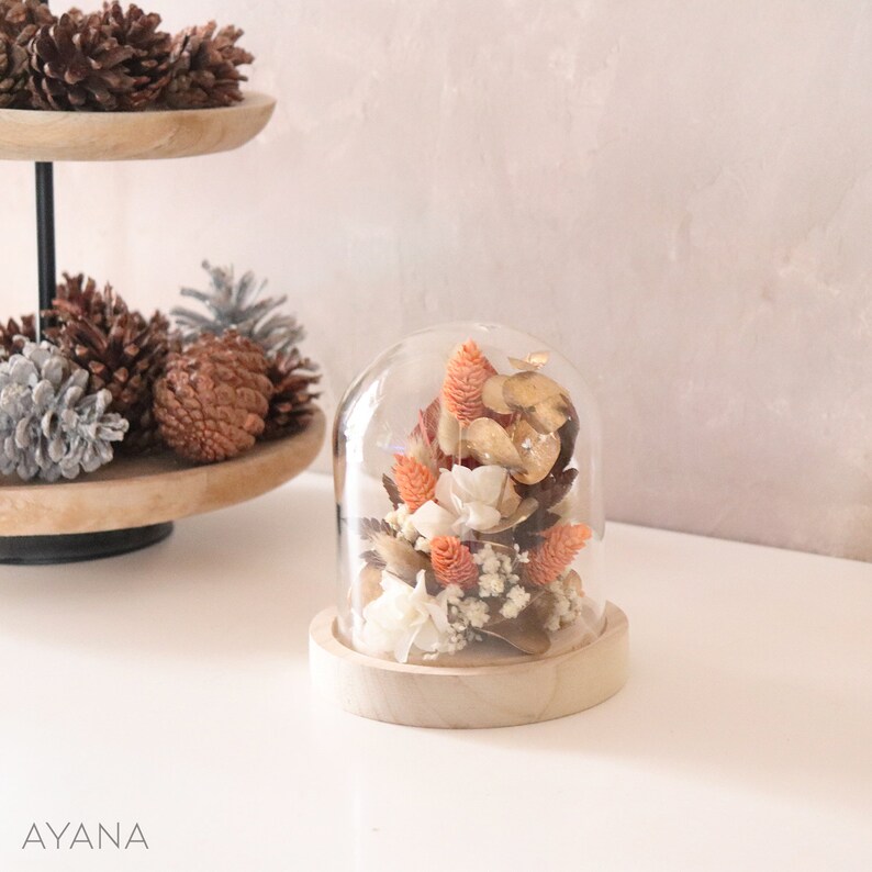 Op de afbeelding: Een glazen stolp met een houten voet die gedroogde bloemen bevat in tinten oranje, wit en bruin. De bloemen zijn in een tros gerangschikt en omvatten kleine witte bloemen, oranje bloemen en bruine bladeren.