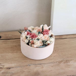 Op de afbeelding: Een ronde, lichtroze container gevuld met gedroogde bloemen. De bloemen zijn een mix van roze, wit en crèmekleuren. De container staat op een houten oppervlak.