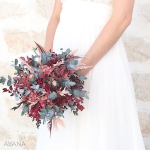 May include: A bridal bouquet with burgundy, blush pink, and white flowers, greenery, and dried grasses. The bouquet is held by a person wearing a white dress.