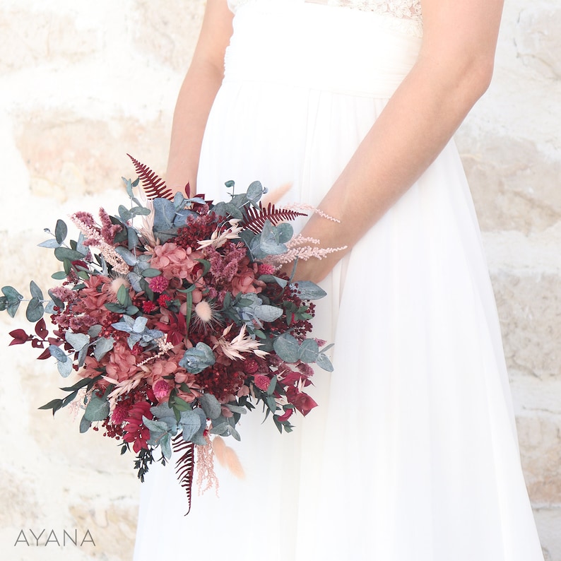 May include: A bridal bouquet with burgundy, blush pink, and white flowers, greenery, and dried grasses. The bouquet is held by a person wearing a white dress.