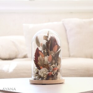 Op de afbeelding: Een glazen stolp met een houten voet, die een gedroogde bloemenarrangement bevat. De bloemen zijn in tinten bruin, beige en oranje.
