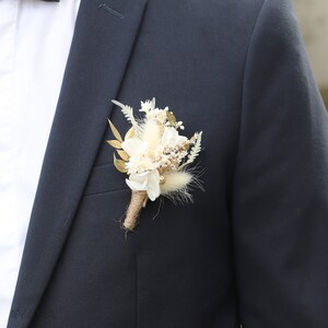 May include: A small bouquet of dried flowers in shades of white and cream, with gold accents, is pinned to a dark blue suit jacket.