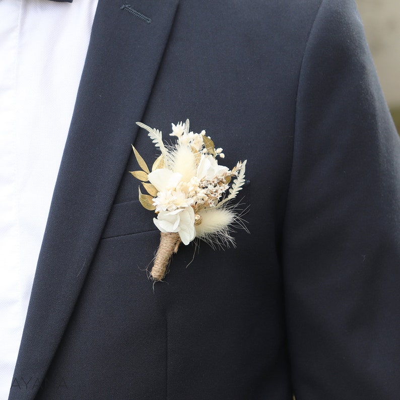 May include: A small bouquet of dried flowers in shades of white and cream, with gold accents, is pinned to a dark blue suit jacket.