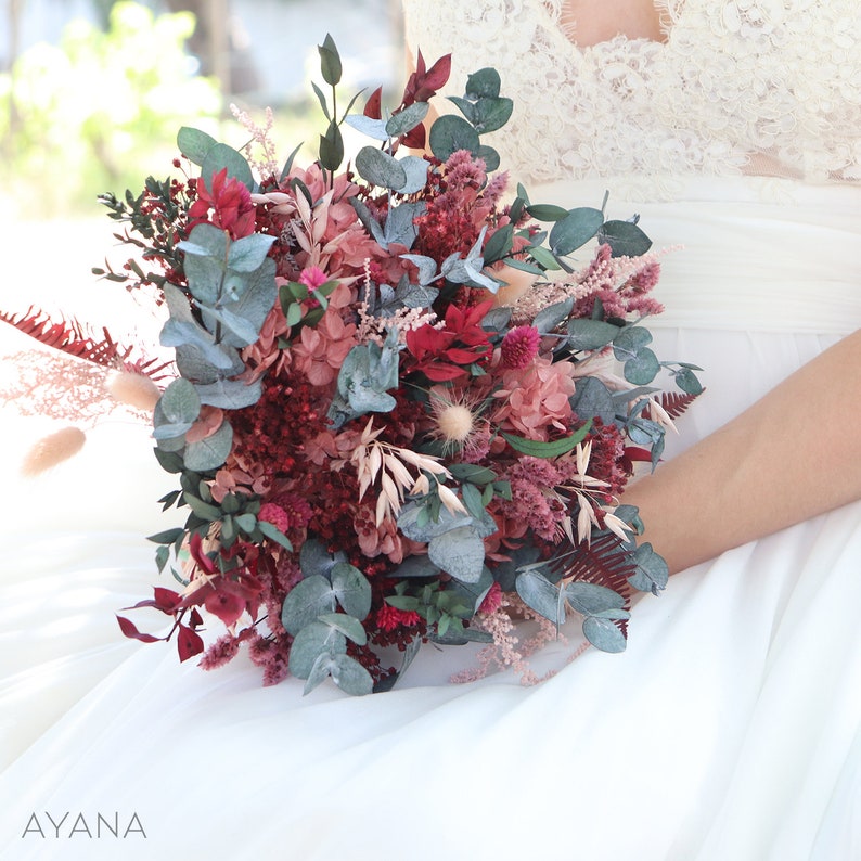 May include: A bridal bouquet with a mix of dried flowers and greenery in shades of burgundy, pink, and green. The bouquet features eucalyptus, dried hydrangeas, and other dried flowers.