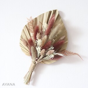 Könnte beinhalten: Ein Trockenblumenarrangement mit einem großen, fächerförmigen Palmenblatt. Der Strauß enthält bordeauxfarbene, beige und weiße Trockenblumen, die mit Bindfaden gebunden sind. Das Wort "AYANA" steht unten.