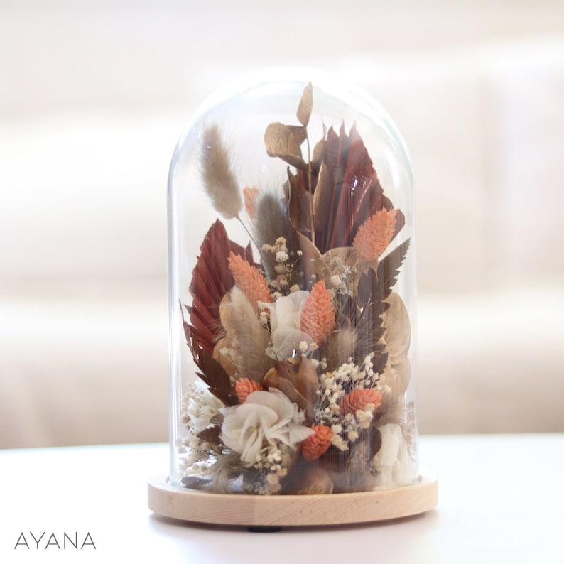 Op de afbeelding: Een glazen stolp met een houten voet die een gedroogde bloemenarrangement bevat. De opstelling bevat gedroogde bloemen in tinten bruin, wit en oranje.