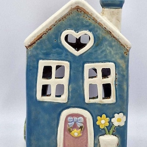 May include: A blue ceramic house-shaped candle holder with a heart-shaped window, two square windows, and a chimney. The house has a pink door with a bow and wreath, and a small white pot with yellow and white flowers.