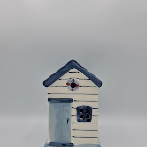 May include: A ceramic beach hut ornament in white and blue. The hut has a blue roof, door, and window frame. A red and white life preserver is painted on the front. The ornament is designed to resemble a seaside structure.