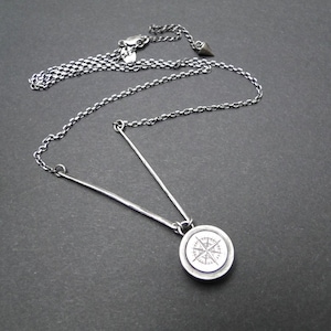 Puede incluir: Un collar de tono plateado oscuro que presenta una barra de metal delgada en forma de V unida a un colgante circular. El colgante muestra un diseño detallado de rosa de los vientos grabado. El collar incluye una cadena delicada.
