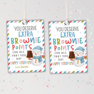 Könnte beinhalten: Zwei rechteckige Geschenkanhänger mit festlichem Design. Jeder Anhänger zeigt den Text "YOU DESERVE EXTRA BROWNIE POINTS" und eine Schneemann-Illustration. Die Anhänger haben einen rot-weiß-blauen Streifenrand und lesen "HAPPY HOLIDAYS!" und "Love, Danisha."