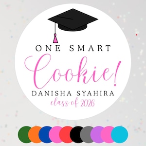 Puede incluir: Una pegatina circular blanca con un birrete negro y una borla rosa. El texto dice "ONE SMART Cookie!" en negro y rosa, con el nombre "DANISHA SYAHIRA" y "class of 2026". Se muestra una paleta de colores debajo.