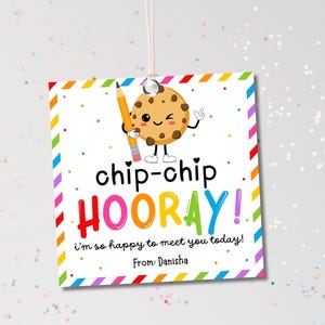 May include: A square gift tag with a cartoon cookie holding a pencil, the words "chip-chip HOORAY!" in colorful letters, and the message "I'm so happy to meet you today!" The tag has a rainbow border and a string.