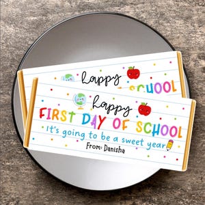 May include: Two chocolate bar wrappers with a school theme. The wrappers have the text "Happy School" and "First Day of School" in colorful letters. The wrappers also have illustrations of an apple, a globe, and a pencil.