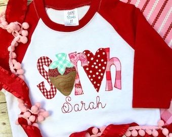 May include: White and red raglan t-shirt with a patchwork design that spells out "love" with a strawberry in the middle. The shirt has the name "Sarah" embroidered below the design.