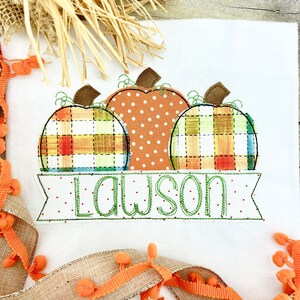 May include: A white fabric with three embroidered pumpkins in orange, green, and yellow plaid. The pumpkins are arranged in a row with the name "Lawson" embroidered in green below them.