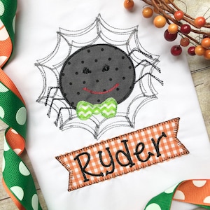 May include: A white onesie with a black spider wearing a green bow tie in a gray spider web. The onesie has a banner with the name "Ryder" in black text on an orange and white checkered background.