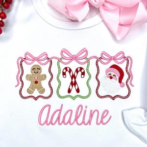 May include: White baby shirt with pink embroidered Christmas designs: a gingerbread man, candy canes, and Santa Claus. The name "Adaline" is embroidered in pink script below the designs. A pink bow is visible in the background.