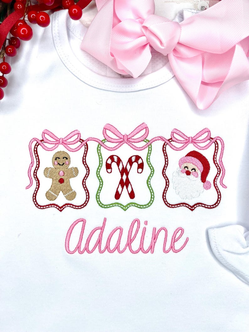 May include: White baby shirt with pink embroidered Christmas designs: a gingerbread man, candy canes, and Santa Claus. The name "Adaline" is embroidered in pink script below the designs. A pink bow is visible in the background.