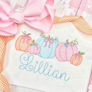 Pu&ograve; includere: Una camicia bianca ricamata con una fila di cinque zucche di diversi colori e il nome "Lillian" ricamato sotto. Le zucche sono arancioni, rosa, blu, rosa e arancioni.