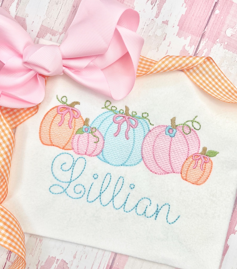 Pu&ograve; includere: Una camicia bianca ricamata con una fila di cinque zucche di diversi colori e il nome "Lillian" ricamato sotto. Le zucche sono arancioni, rosa, blu, rosa e arancioni.