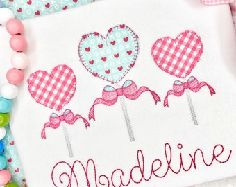 May include: A white onesie with three embroidered pink and blue heart-shaped lollipops and the name "Madeline" in pink cursive.