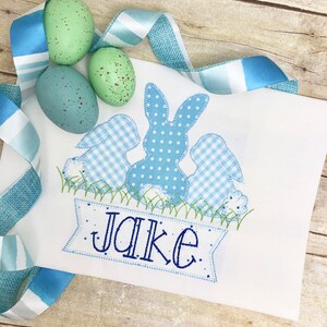 May include: A white embroidered appliqu&eacute; with three blue bunnies, one with polka dots and two with gingham patterns. The bunnies are sitting in a green grass patch with a blue banner that reads "Jake".