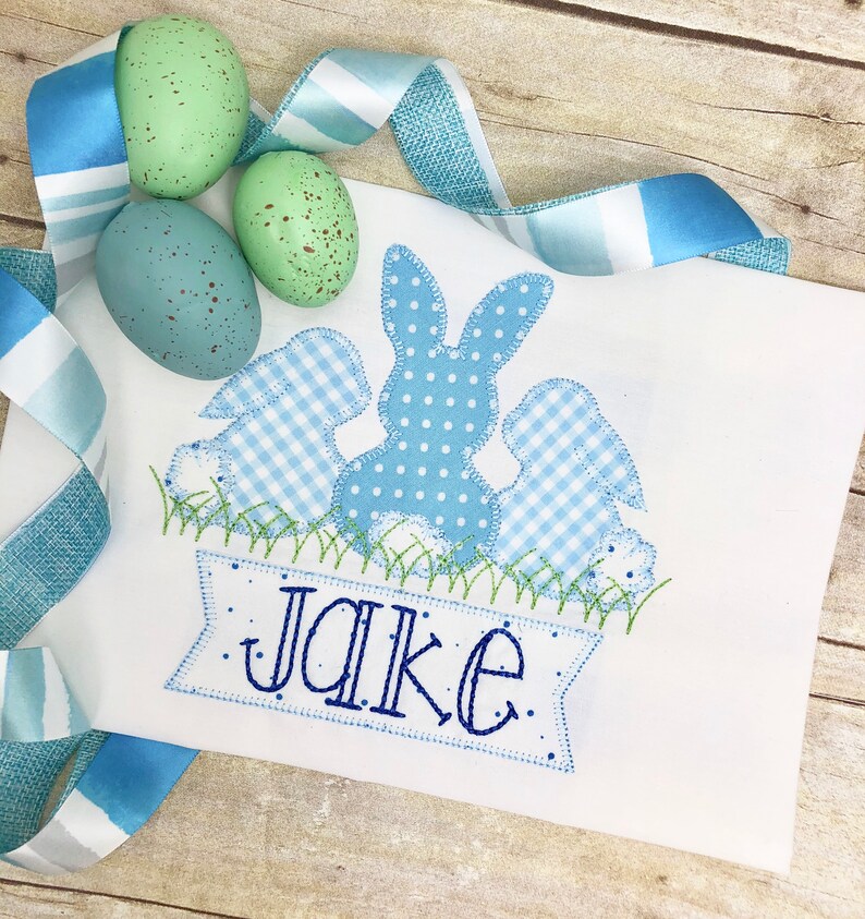 May include: A white embroidered appliqu&eacute; with three blue bunnies, one with polka dots and two with gingham patterns. The bunnies are sitting in a green grass patch with a blue banner that reads "Jake".