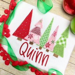 Puede incluir: Camiseta blanca con un dise&ntilde;o de patchwork de &aacute;rboles de Navidad en rojo, rosa y verde. El nombre "Quinn" est&aacute; bordado en rojo debajo de los &aacute;rboles.