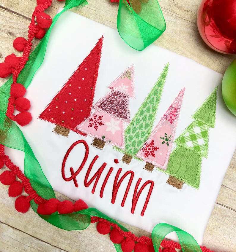 Puede incluir: Camiseta blanca con un dise&ntilde;o de patchwork de &aacute;rboles de Navidad en rojo, rosa y verde. El nombre "Quinn" est&aacute; bordado en rojo debajo de los &aacute;rboles.