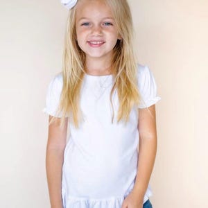 Pu&ograve; includere: Una bambina con lunghi capelli biondi indossa una camicia bianca a maniche corte con un bordo a balze. Sorride e ha un fiocco bianco nei capelli.