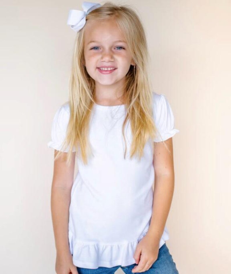 Pu&ograve; includere: Una bambina con lunghi capelli biondi indossa una camicia bianca a maniche corte con un bordo a balze. Sorride e ha un fiocco bianco nei capelli.