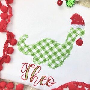 May include: A white onesie with a green and white gingham dinosaur wearing a red Santa hat. The dinosaur is holding a red ornament. The onesie is personalized with the name "Theo" in green and red embroidery.