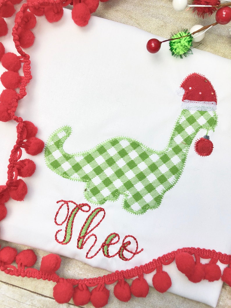 May include: A white onesie with a green and white gingham dinosaur wearing a red Santa hat. The dinosaur is holding a red ornament. The onesie is personalized with the name "Theo" in green and red embroidery.