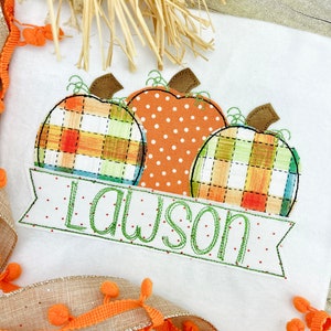 May include: A white t-shirt with an embroidered design of three pumpkins. The pumpkins are in a plaid pattern, a polka dot pattern, and a plaid pattern. The text "Lawson" is embroidered on a banner below the pumpkins.