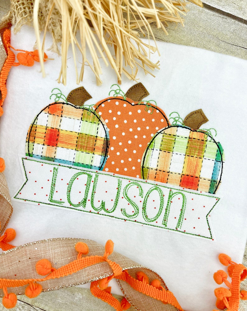 May include: A white t-shirt with an embroidered design of three pumpkins. The pumpkins are in a plaid pattern, a polka dot pattern, and a plaid pattern. The text "Lawson" is embroidered on a banner below the pumpkins.