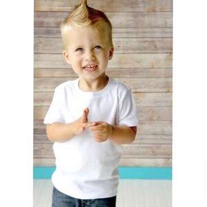 May include: A young boy with a spiky blond hairstyle is wearing a white t-shirt. He is smiling and looking up.