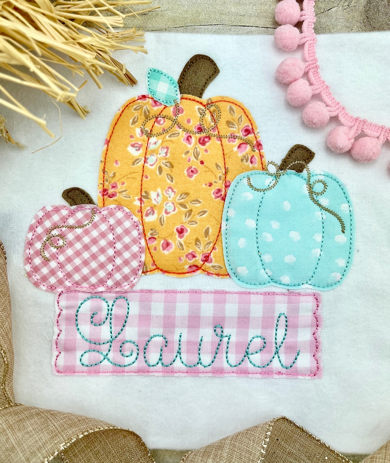 May include: A white fabric with three embroidered pumpkins. The pumpkins are in pink, yellow, and blue with floral patterns. The fabric has the name "Laurel" embroidered in pink on a pink and white gingham background.