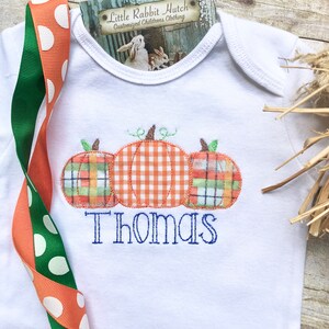 May include: White baby onesie with three embroidered pumpkins in orange, green, and plaid. The pumpkins are stacked on top of each other. The name "Thomas" is embroidered below the pumpkins in blue thread.