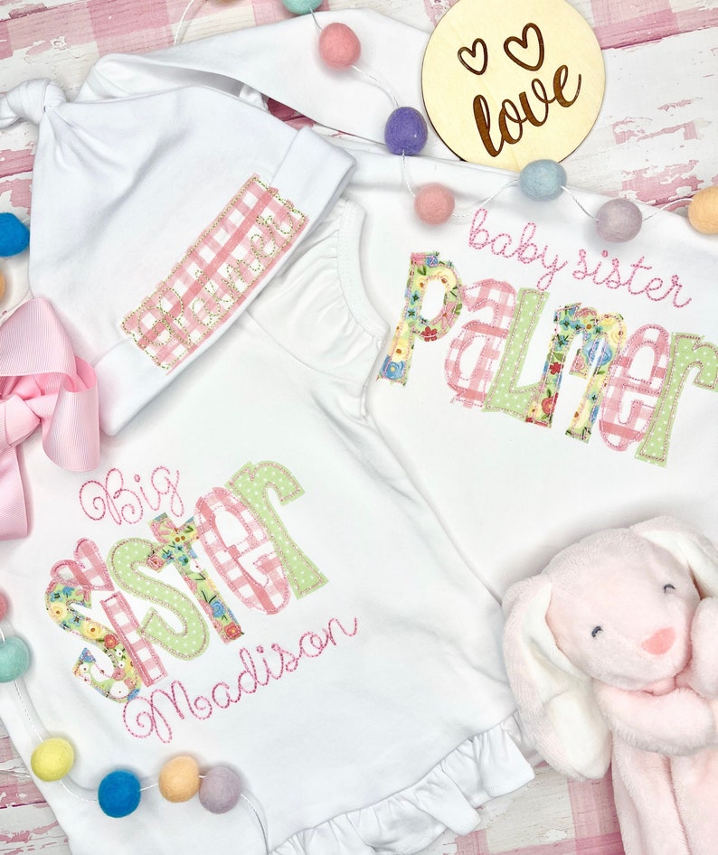May include: Two white babygrows with pink and green floral and gingham designs. The larger babygrow says "Big Sister Madison" and the smaller babygrow says "Baby Sister Palmer".