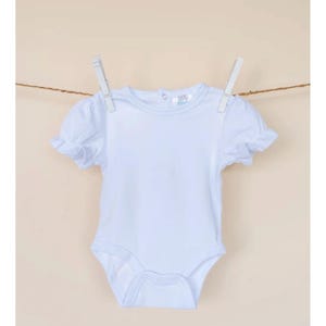 Pu&ograve; includere: Un body bianco per neonato con maniche corte e un bordo a balze. Il body ha una scollatura rotonda e una chiusura a pressione sul fondo.