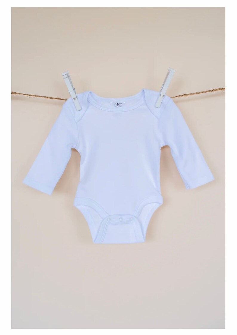 May include: A white long-sleeved baby onesie hanging on a clothesline with two clothespins. The onesie has a small logo on the front that says "6-12M".
