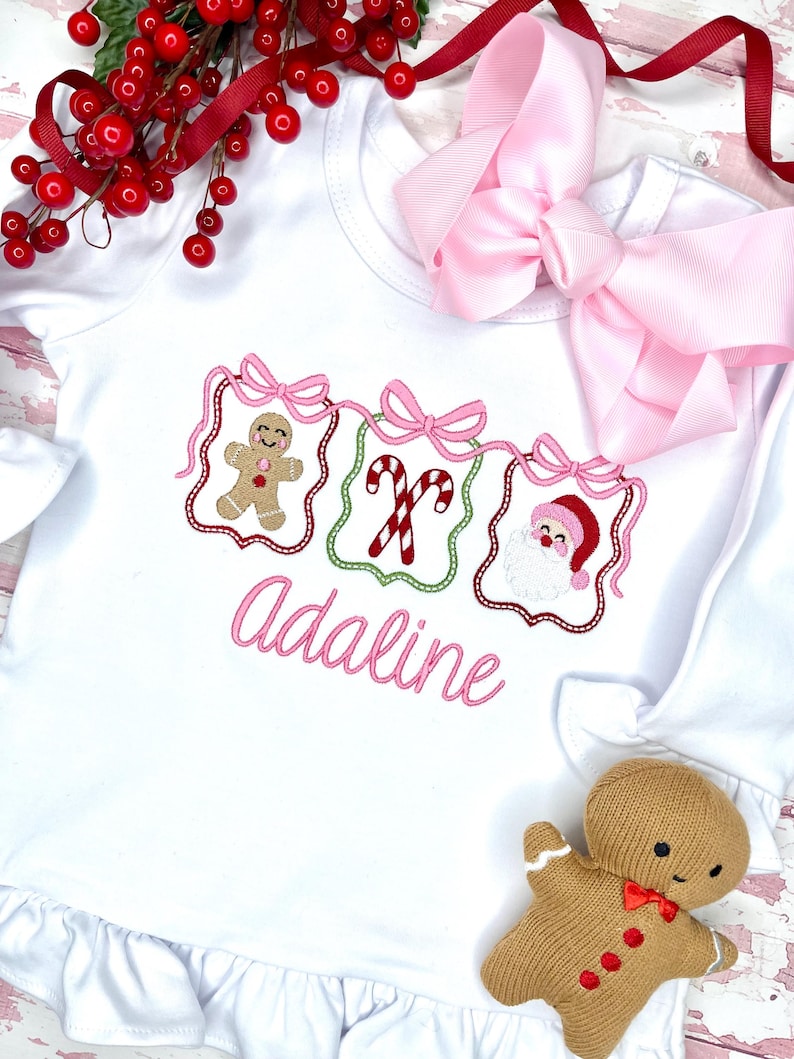 May include: White long-sleeved baby shirt with a ruffle hem, featuring embroidered Christmas designs: a gingerbread man, candy canes, and Santa Claus. The name "Adaline" is embroidered in pink. A large pink bow and a gingerbread doll are also present.