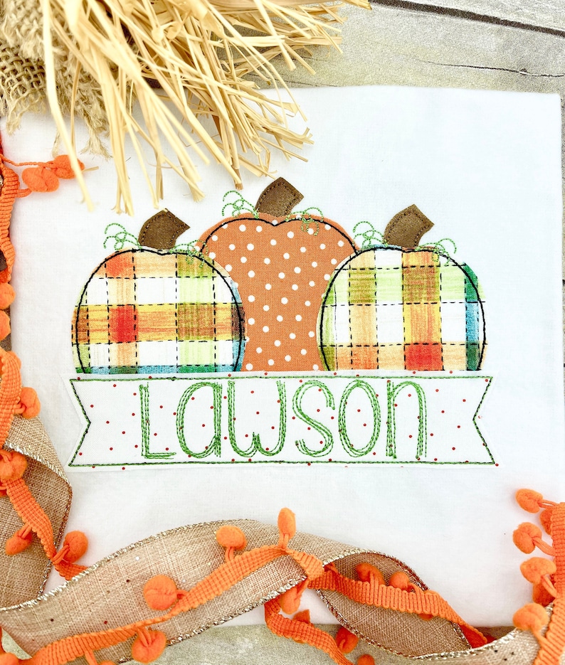 May include: A white fabric with three embroidered pumpkins in orange, green, and yellow plaid. The pumpkins are arranged in a row with the name "Lawson" embroidered in green below them.