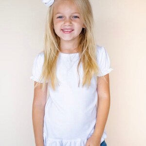 May include: A young girl with long blonde hair wearing a white short-sleeved shirt with a ruffle trim. She is smiling and has a white bow in her hair.