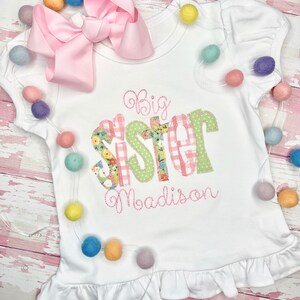 May include: White t-shirt with a pink bow on top. The shirt has a green and pink patchwork design that says "Big Sister" with the name "Madison" embroidered underneath.